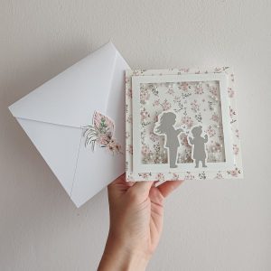 Čestitka za krštenje dve sestre, sa cvetnim dezenom i siluetama devojčica, uz belu kovertu sa floralnim ukrasom.